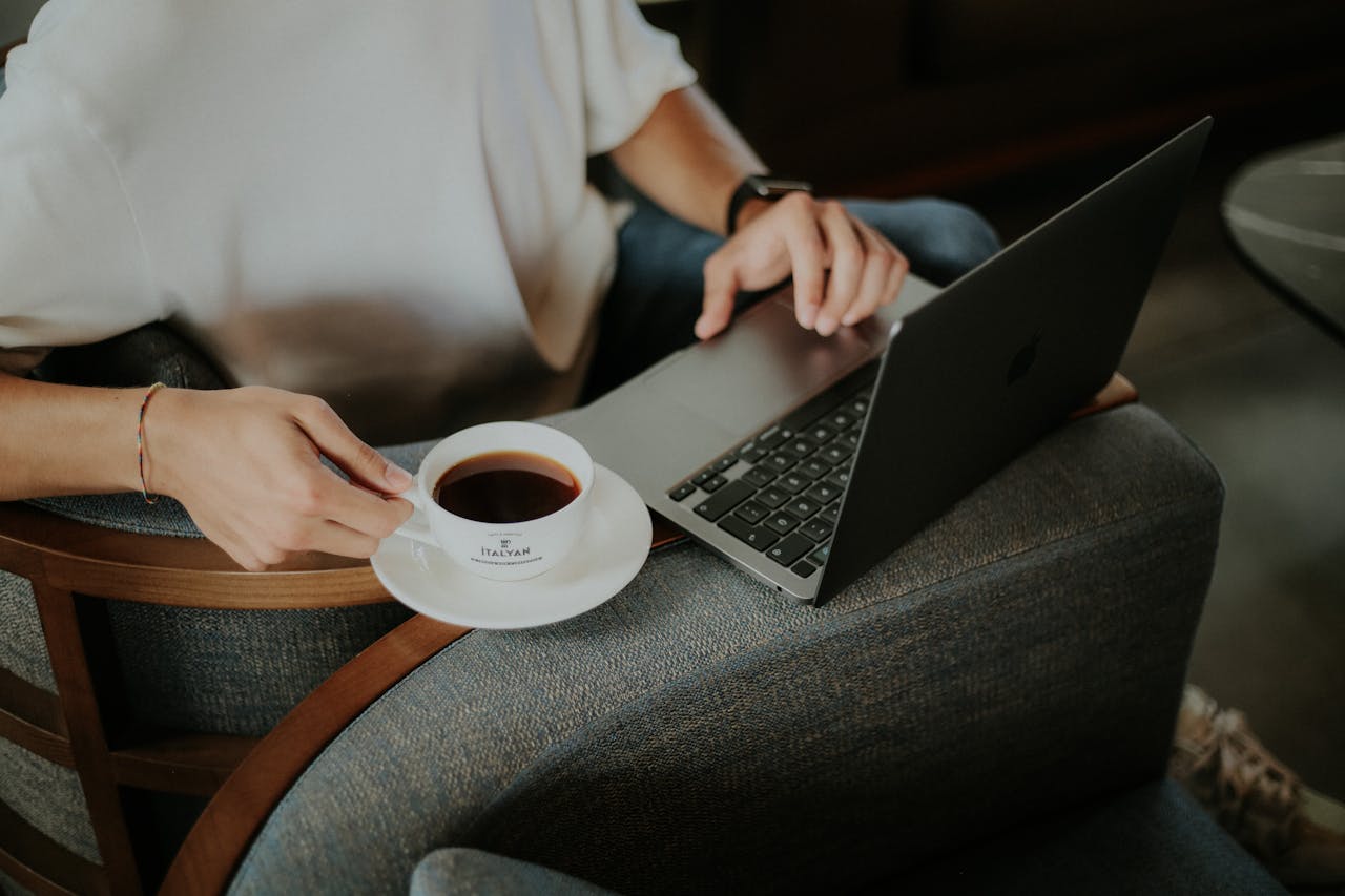 Person enjoying a cup of coffee while using a laptop inside a cozy setting, perfect for work or leisure.